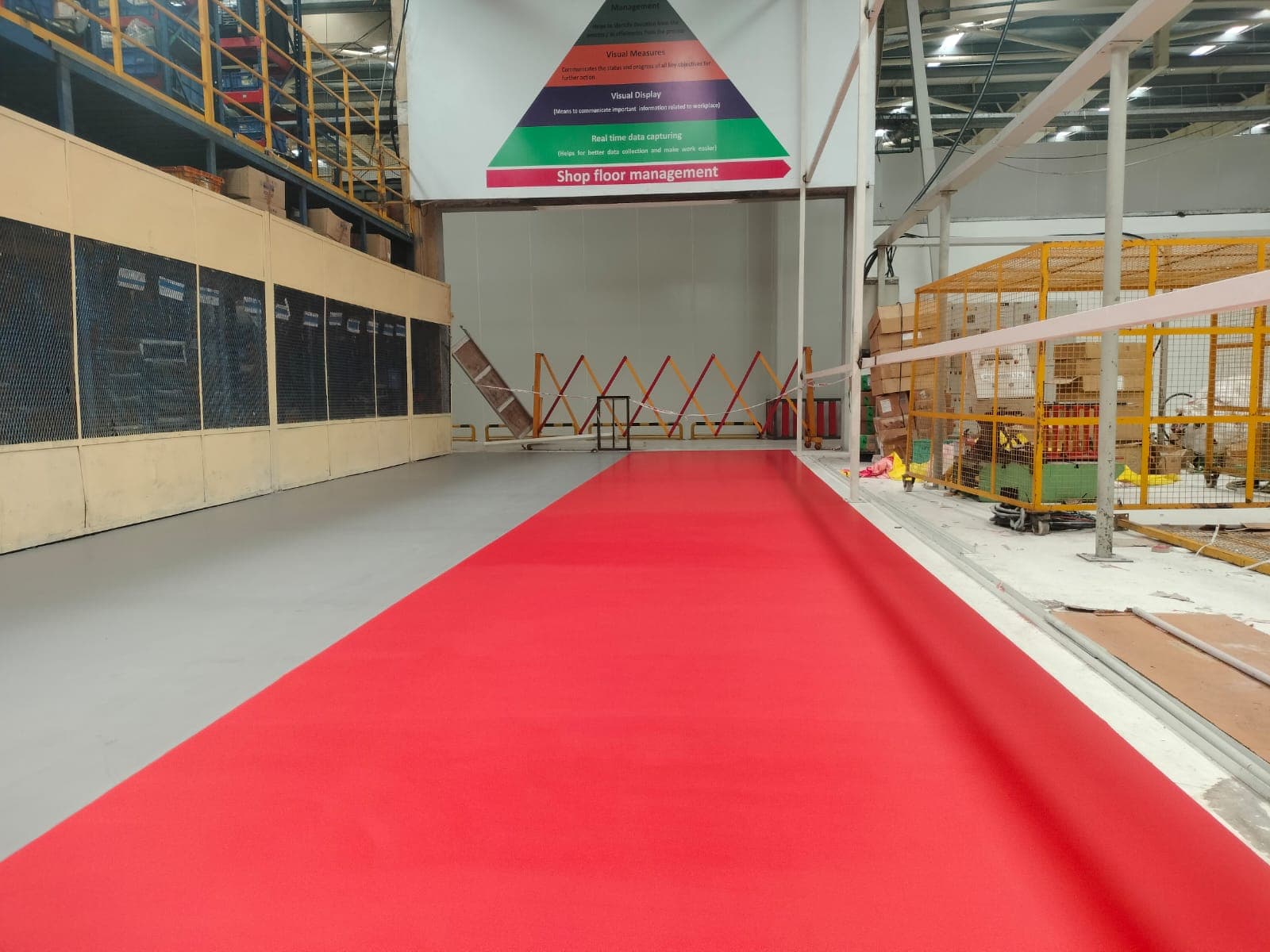 A red epoxy floor walkway inside an industrial warehouse, running between storage racks and a shop-floor-management signage board.